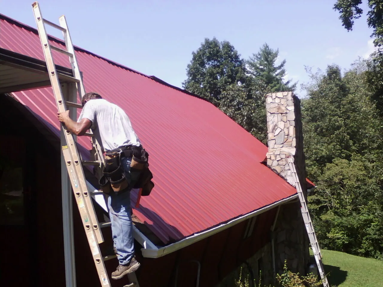 Roofer on a ladder beside a red metal roof and stone chimney — Soffit & Fascia Repair by Roofing Brothers in Grass Valley